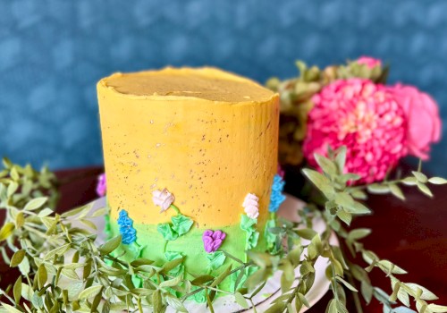 A tall cake decorated with yellow and green frosting, featuring small floral designs, surrounded by leafy garlands and pink flowers on a table.