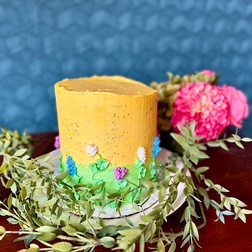 A tall cake decorated with yellow and green frosting, featuring small floral designs, surrounded by leafy garlands and pink flowers on a table.