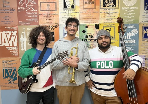 Three men are posing with musical instruments in front of a wall of jazz posters. One holds a guitar, the second a trumpet, and the third a double bass.