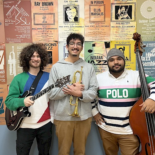 Three men are posing with musical instruments in front of a wall of jazz posters. One holds a guitar, the second a trumpet, and the third a double bass.