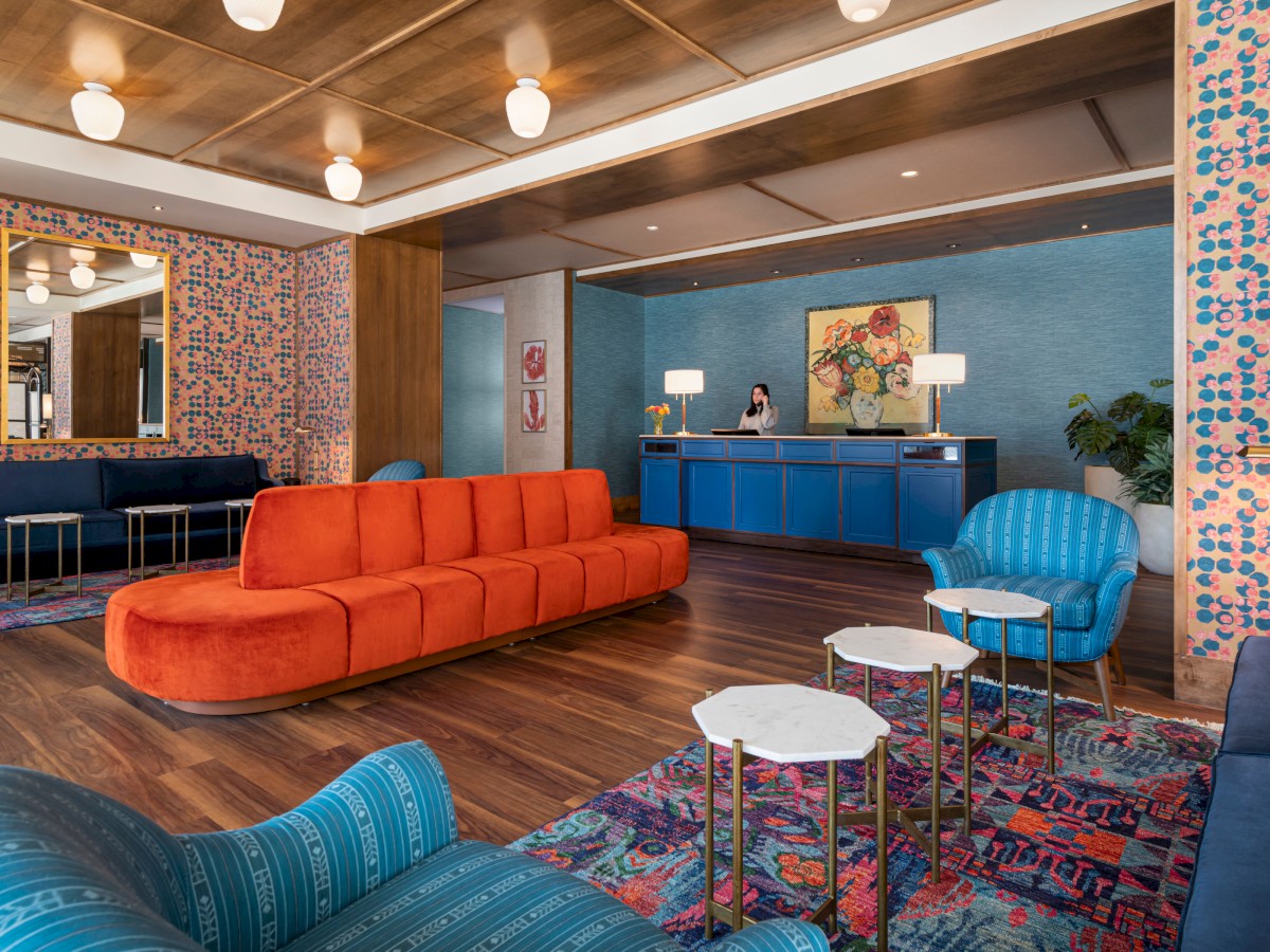 A modern hotel lobby features a long orange sofa, blue armchairs, patterned wallpaper, colorful rugs, and a reception desk with a friendly staff member.