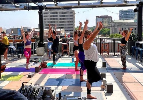 People are practicing yoga on a rooftop, with a DJ setup in the foreground and a cityscape in the background.