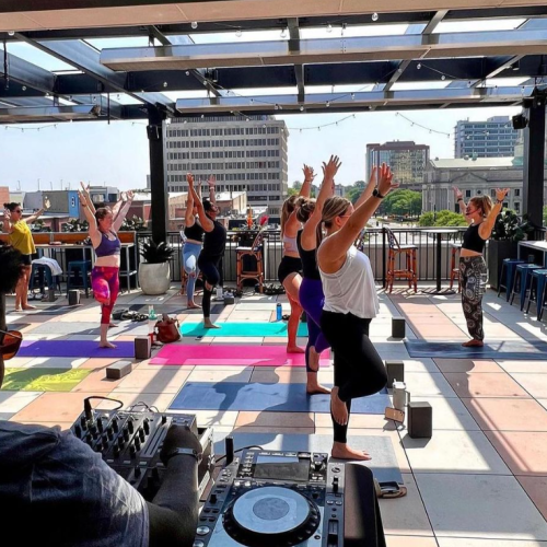 People are practicing yoga on a rooftop, with a DJ setup in the foreground and a cityscape in the background.