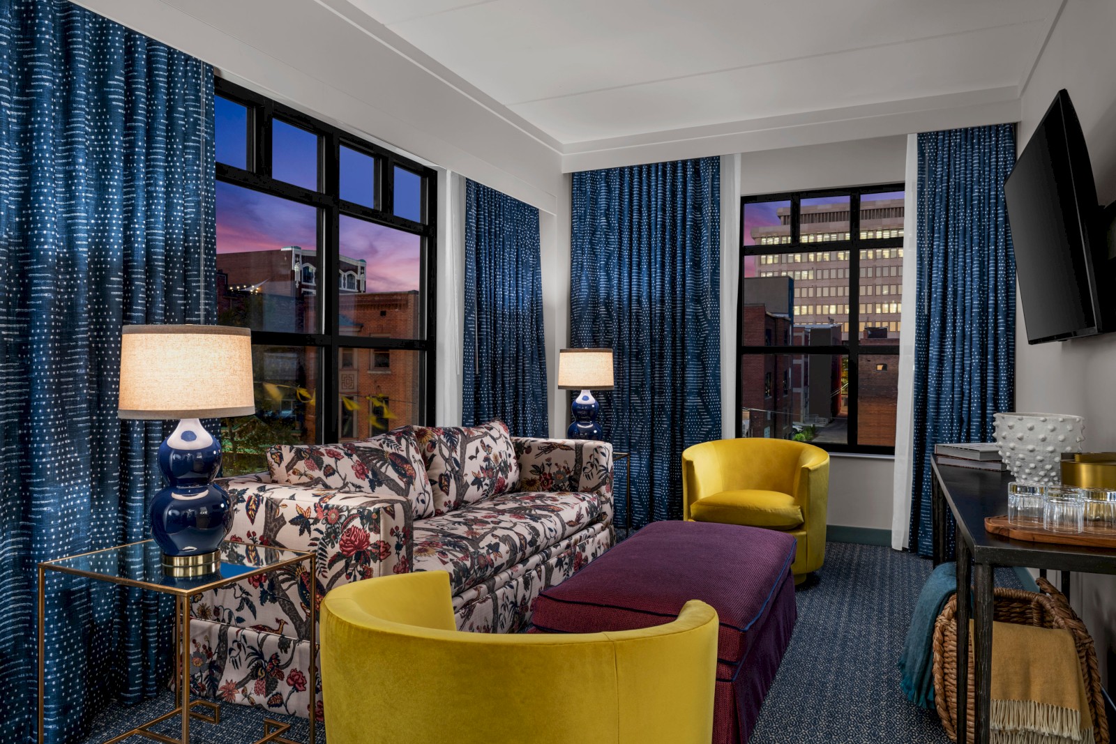 A chic living room with a floral sofa, two yellow chairs, blue curtains, and two table lamps, featuring large windows overlooking a cityscape.