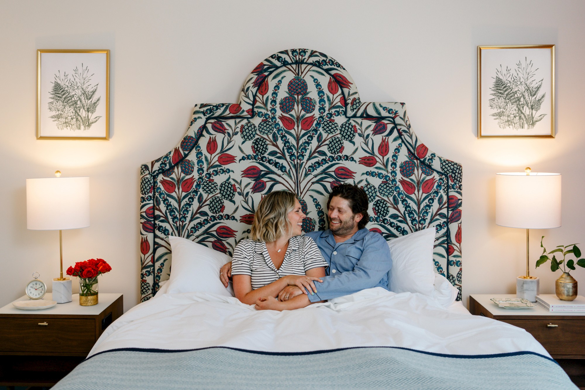 A couple sits smiling in bed with a floral headboard, flanked by lamps, framed art, nightstands, and decor.