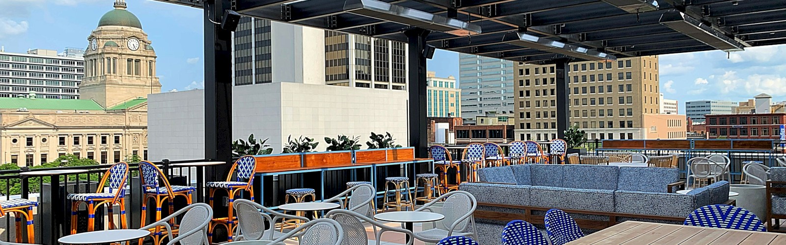 Outdoor rooftop seating area with tables and chairs, featuring city skyline views and nearby buildings, partially covered by a pergola structure.