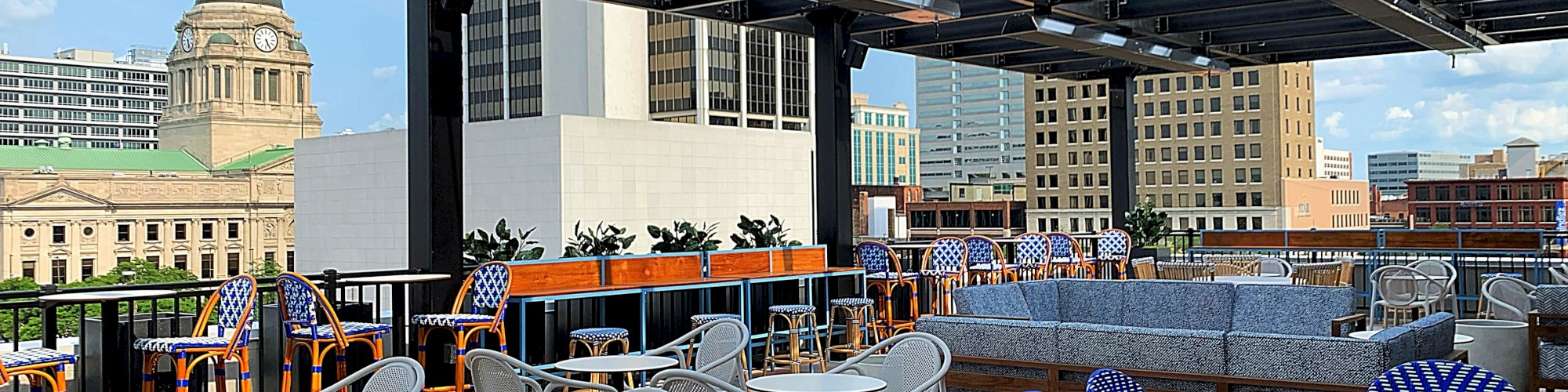 Outdoor rooftop seating area with tables and chairs, featuring city skyline views and nearby buildings, partially covered by a pergola structure.