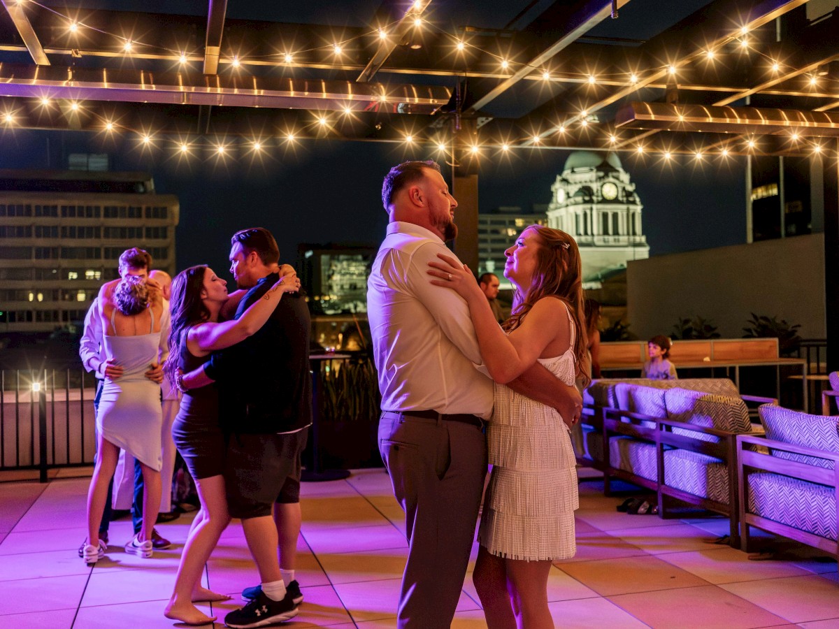 People dancing under string lights on a rooftop terrace at night, with a cityscape in the background.