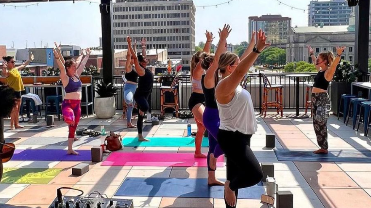 A group of people is practicing yoga on a rooftop terrace with colorful mats, while a DJ plays music in the foreground.