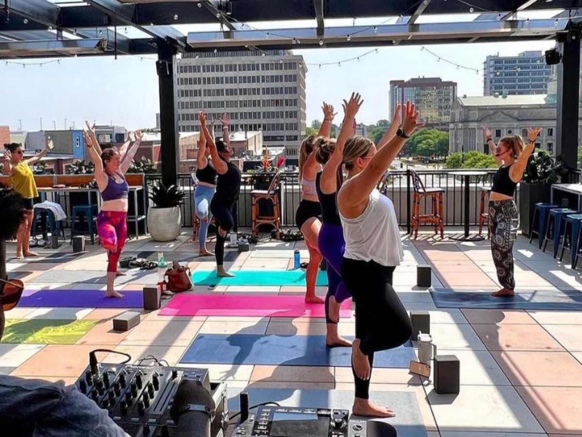 A group of people is practicing yoga on a rooftop terrace with colorful mats, while a DJ plays music in the foreground.