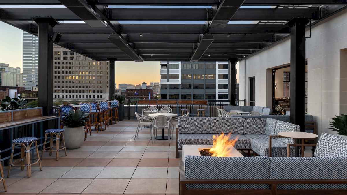 This image shows a modern rooftop patio with seating areas, a fire pit, and bar stools against a cityscape backdrop at sunset.