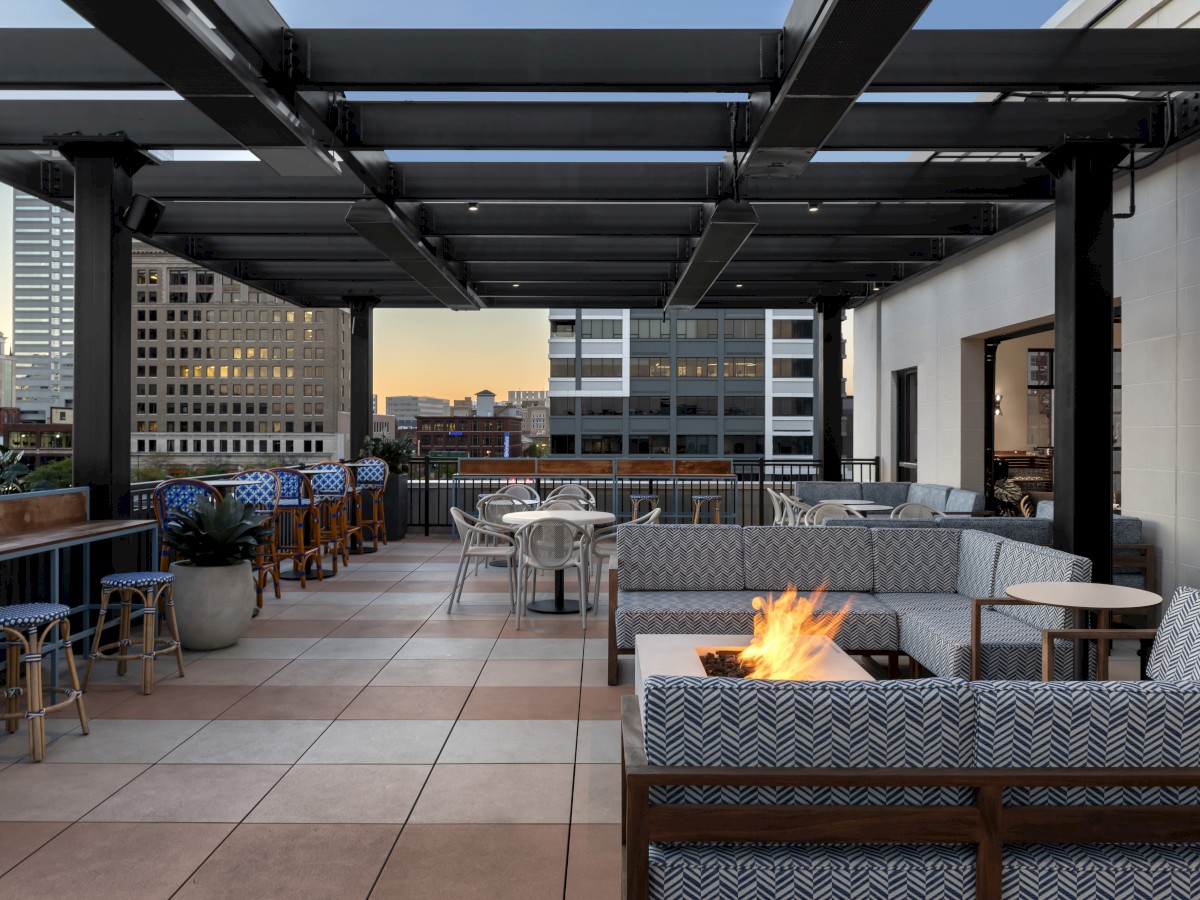 This image shows a modern rooftop patio with seating areas, a fire pit, and bar stools against a cityscape backdrop at sunset.