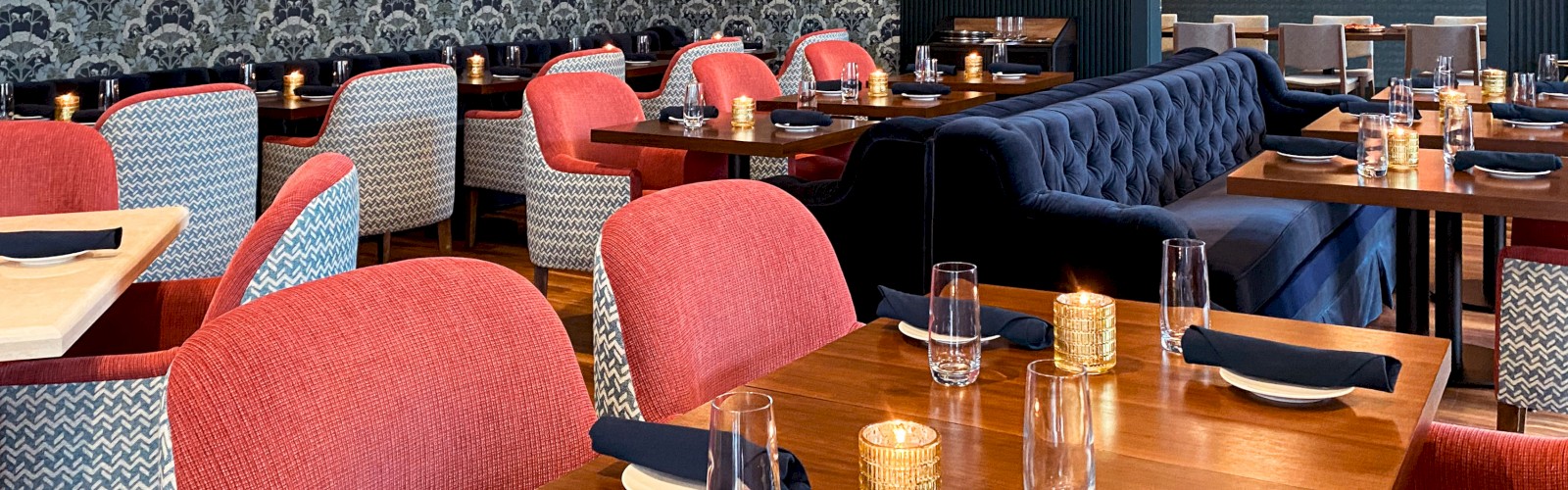 A stylish café with wooden tables, red and blue chairs, hanging lights, blue-patterned wallpaper, and abstract paintings on the walls.