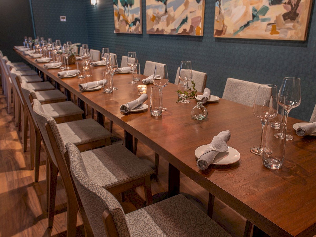 A long dining table is neatly set with glasses, plates, and napkins, surrounded by chairs, in a modern room with abstract paintings on the wall.