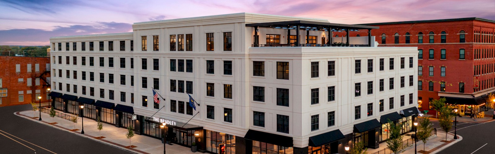 A multi-story white building on a street corner at dusk with outdoor seating and flags displayed, surrounded by an intersection and additional buildings.