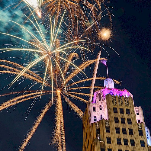 Fireworks burst in the night sky above a lit-up building.