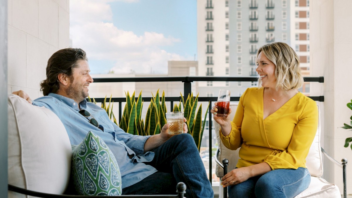 A man and a woman relax with drinks on a balcony, enjoying a sunny day with a cityscape in the background, sitting on comfortable furniture.