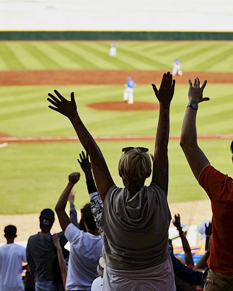 People in a stadium, raising their hands in excitement while watching a baseball game in progress.