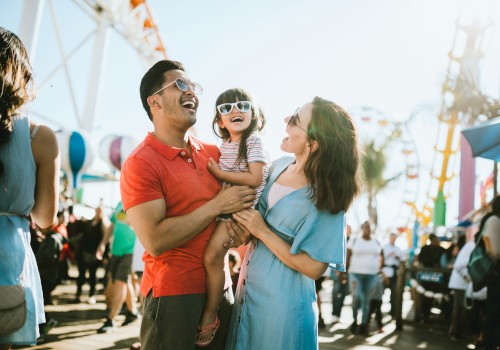 A family of three, with the parents smiling and holding a child in sunglasses, enjoying a sunny day at an amusement park or fair in a crowded area.