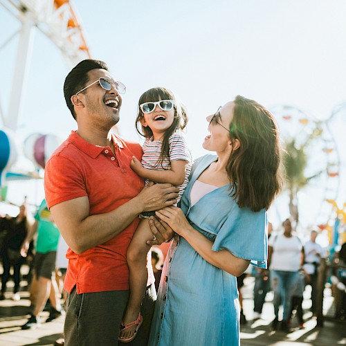 A family of three, with the parents smiling and holding a child in sunglasses, enjoying a sunny day at an amusement park or fair in a crowded area.