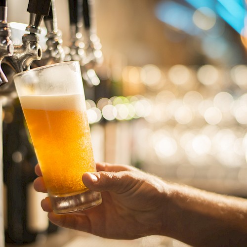 A hand holding a glass of freshly poured beer near a row of taps in a dimly lit bar or pub.