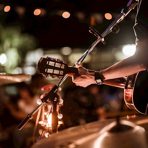 A person is playing an acoustic guitar on stage, with drum equipment visible in the foreground, and a blurred audience in the background.