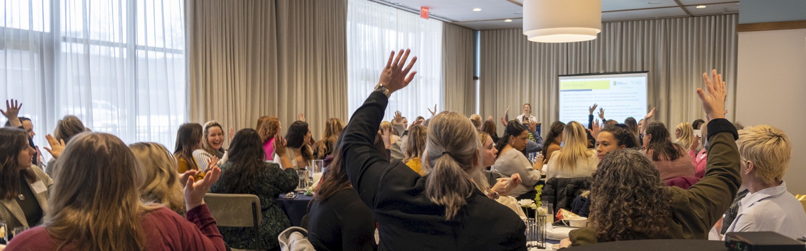 A group of people are seated in a conference room, some with their hands raised, while a speaker presents in front, near a projection screen.