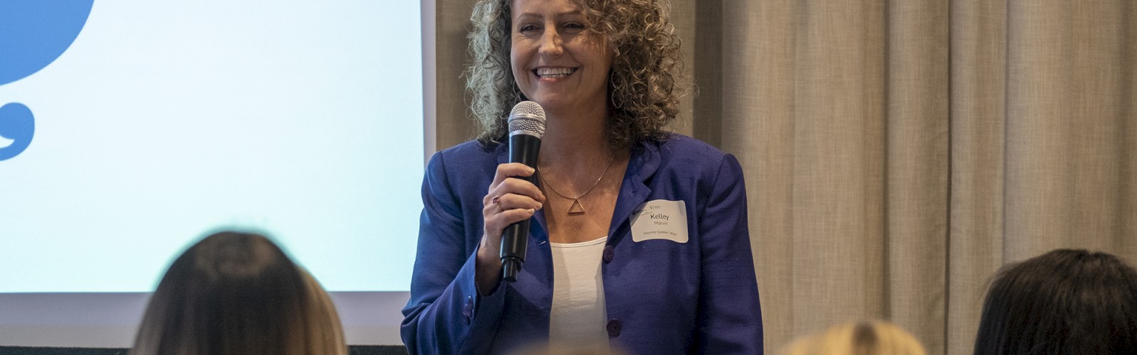 A person is speaking into a microphone in front of a group, with a presentation screen in the background displaying a blue graphic.