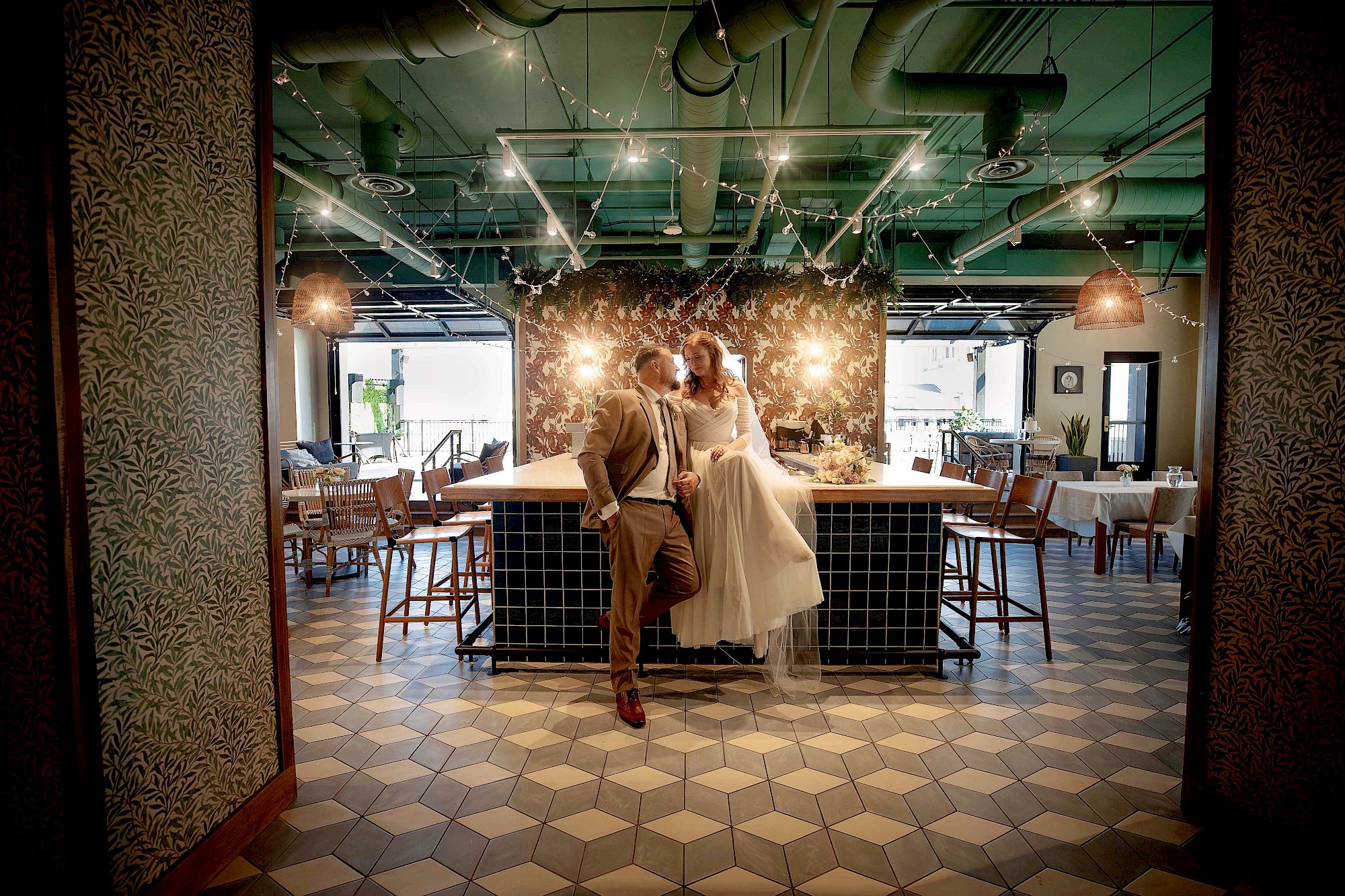 A couple in wedding attire leans against a counter in a stylish, modern restaurant with string lights and patterned tiles adorning the space.