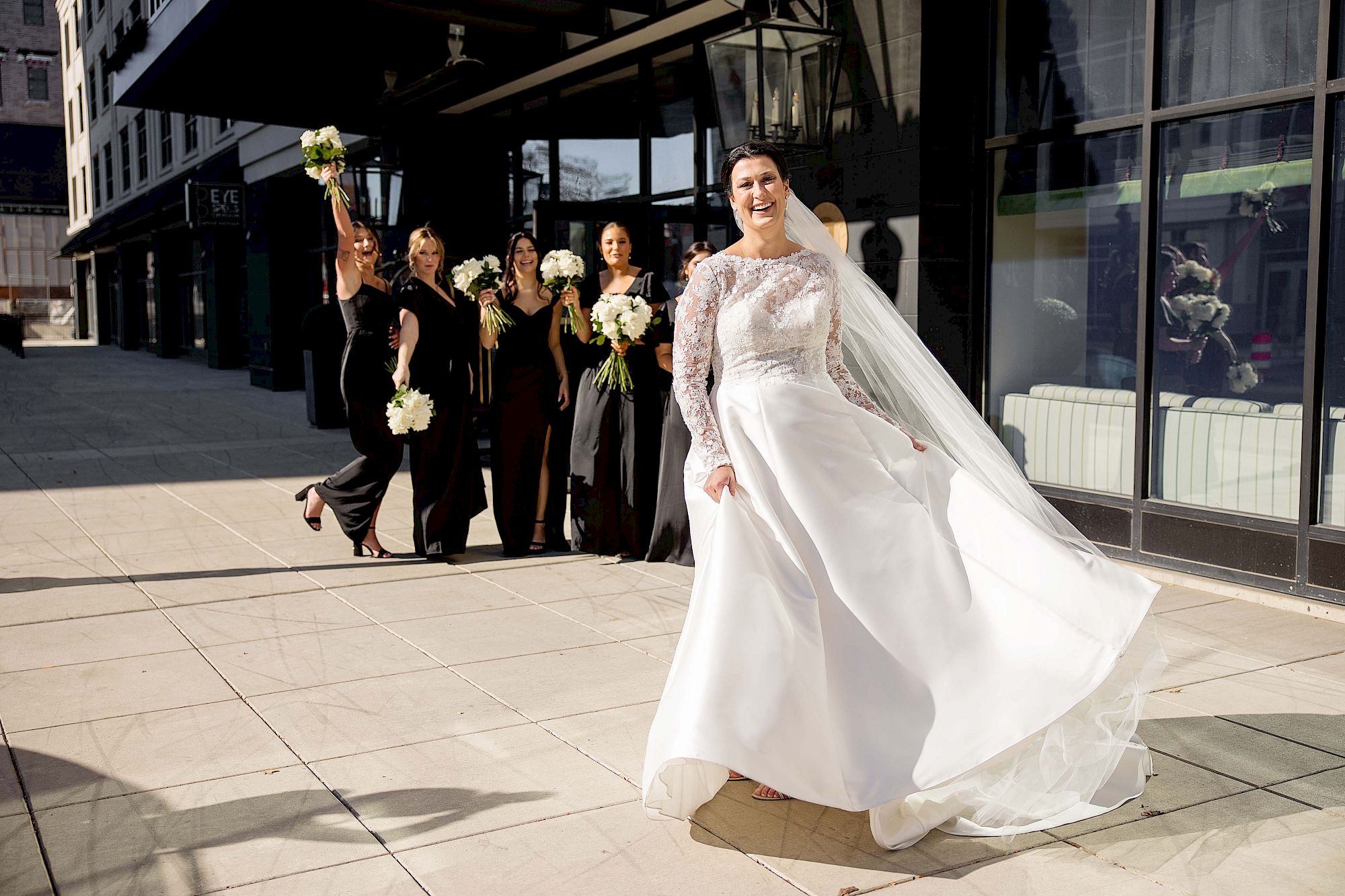 A joyful bride is twirling in her wedding dress outdoors with her bridesmaids cheering and holding bouquets in the background.