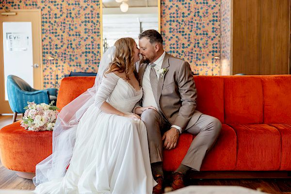 A bride in a white gown and groom in a grey suit share a loving moment on a red couch, sitting close and gazing into each other's eyes.