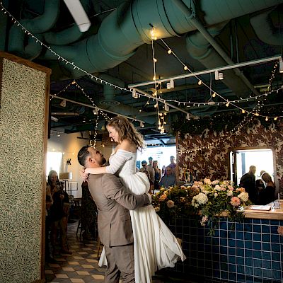 A couple sharing a dance at what appears to be a wedding reception, surrounded by guests, flowers, and festive decorations in a warmly lit venue.