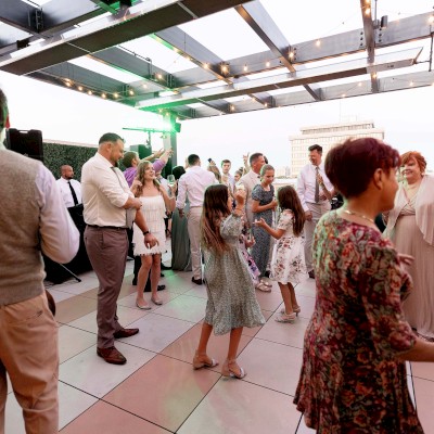 People dancing and socializing at an outdoor event under string lights on a terrace with city views in the background.