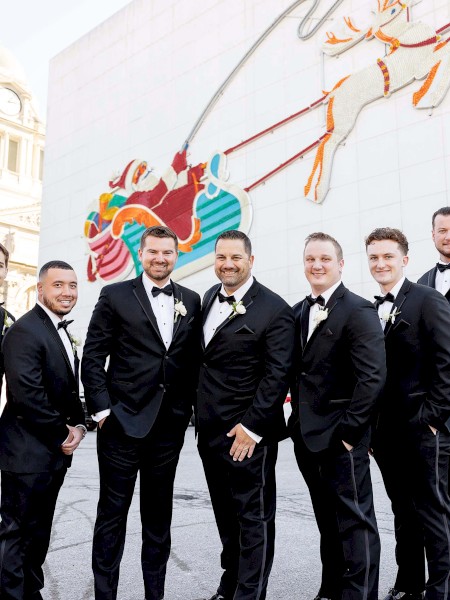 A group of men in black tuxedos stand together outdoors, posing for a photo with a festive mural in the background.