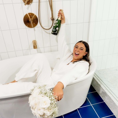 A person in white, holding a champagne bottle and flowers, sits joyfully in a bathtub with a white tiled wall and blue floor tiles in a modern bathroom.