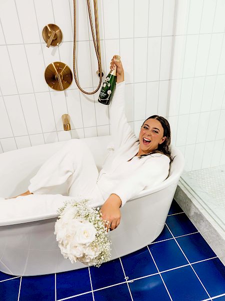 A person in white, holding a champagne bottle and flowers, sits joyfully in a bathtub with a white tiled wall and blue floor tiles in a modern bathroom.