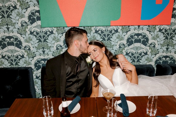 A couple is sitting together, with the man kissing the woman's forehead, surrounded by a patterned wallpaper and table setting.