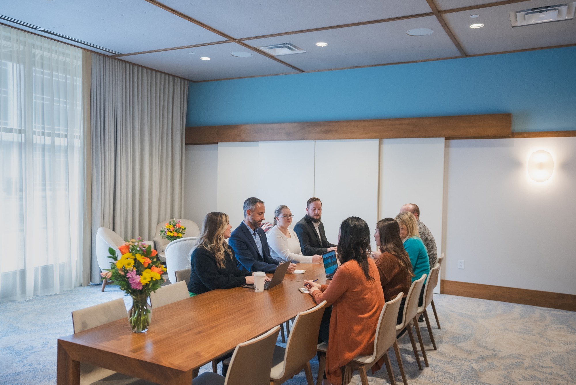 A group of people sitting around a conference table in a well-lit room with flowers on the table, engaged in a meeting or discussion.