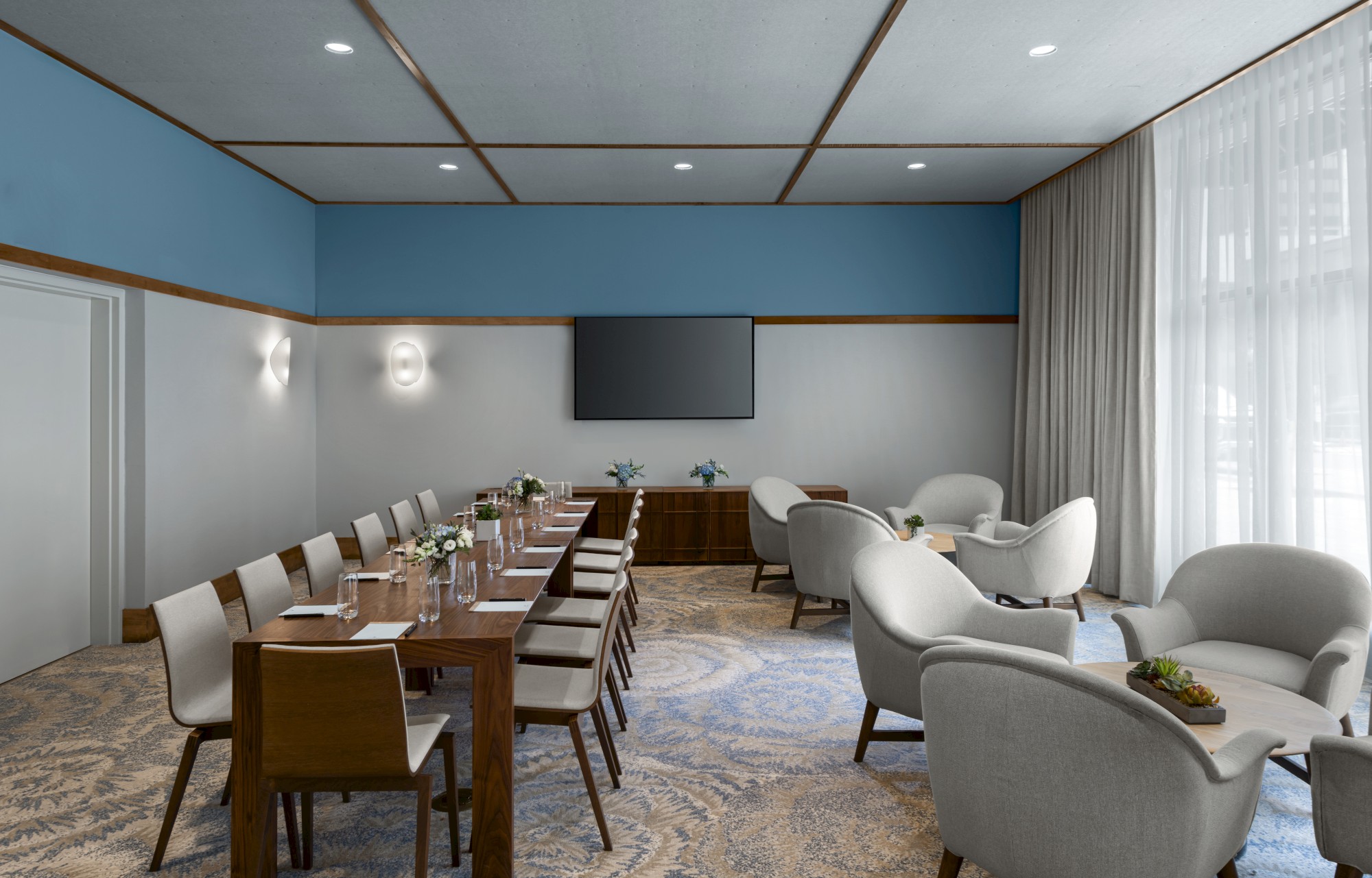 A modern conference room with a long table, chairs, lounge seating, a TV, and decorative plants. Neutral colors and natural light enhance it.