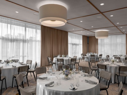 A banquet hall with round tables set for an event, featuring white tablecloths, folded napkins, glassware, and simple centerpieces.