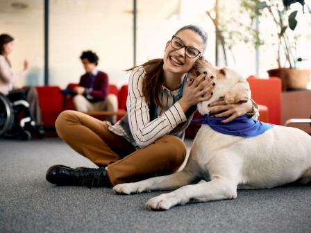 A woman sitting on the floor embraces a large dog while two other people converse in the background of a bright, modern room.