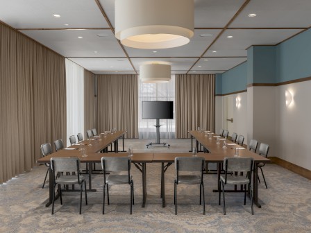 It's a modern conference room with a U-shaped table setup, chairs, a flat-screen TV, and elegant lighting and curtains.