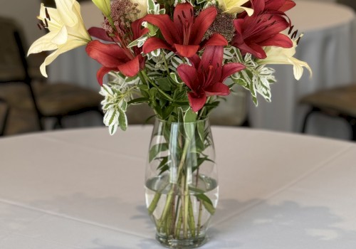 A clear glass vase holds a bouquet of red and white flowers, placed on a white tablecloth-covered table in a room with chairs in the background.