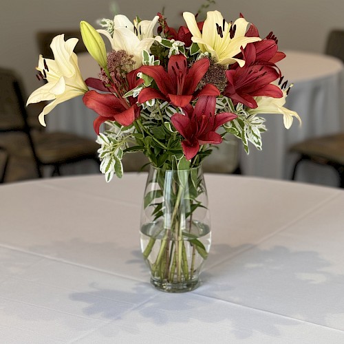 A clear glass vase holds a bouquet of red and white flowers, placed on a white tablecloth-covered table in a room with chairs in the background.