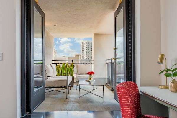 A modern balcony with outdoor seating, green plants, a small table with flowers, and a view of buildings against a blue sky.
