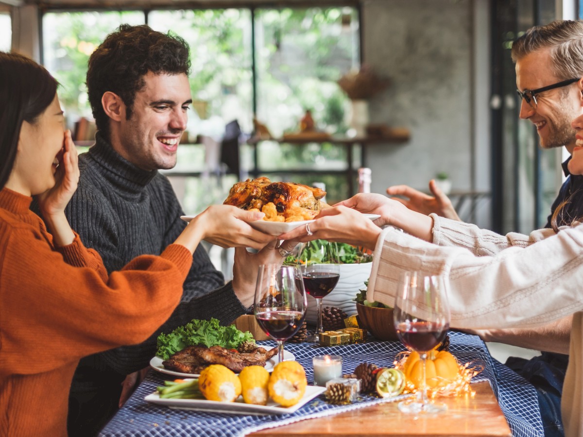 A group of people is gathered around a table, sharing a meal with roast chicken, vegetables, and glasses of wine, enjoying each other's company.