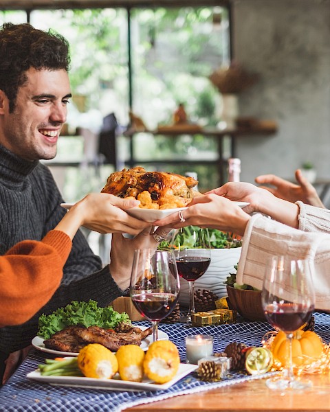 Four people are gathered around a table, smiling and passing a plate of food. The table is set with a meal and wine glasses.