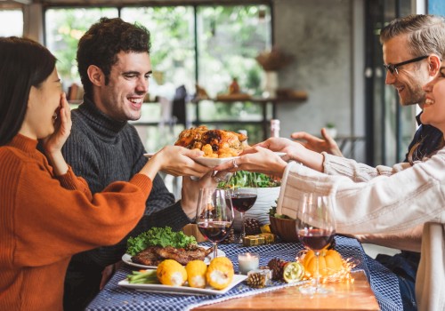 Four people are gathered around a table, smiling and passing a plate of food. The table is set with a meal and wine glasses.