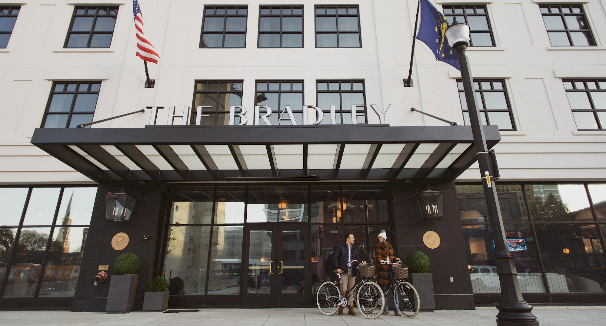 The image shows the facade of The Bradley hotel, with flags and people with bicycles at the entrance.