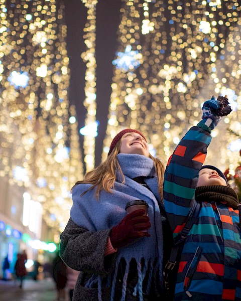 Two people enjoying festive street decorations with warm drinks, surrounded by lights and holiday atmosphere, looking up joyfully.